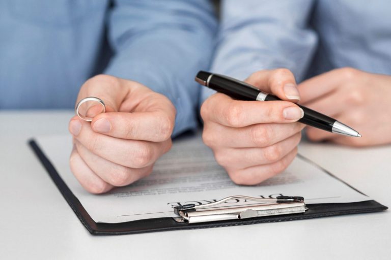 divorce-lawyer-FP Closeup of couple holding wedding ring and pen illustrates blog "How To Choose the Right Divorce Attorney"